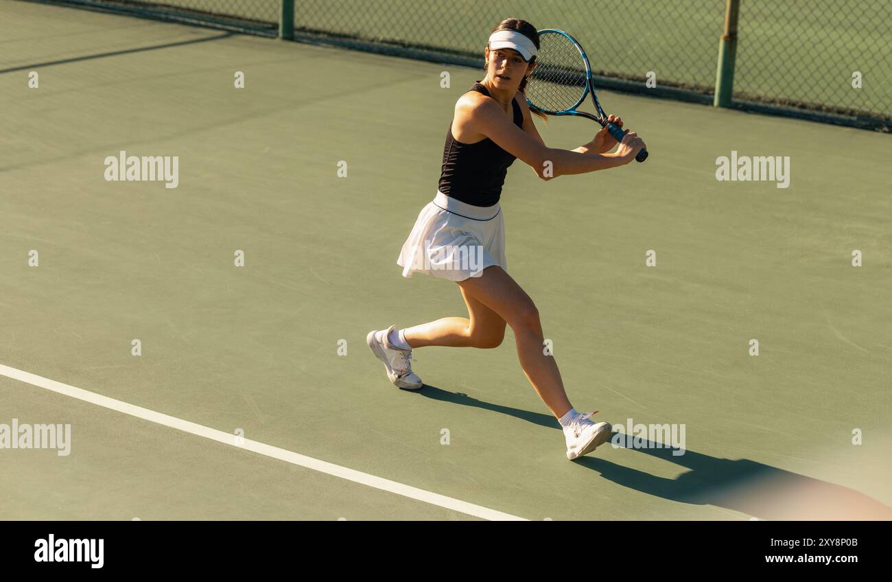 Donna che gioca a tennis, fa oscillare attivamente la sua racchetta su un campo. È vestita con una gonna bianca e una canotta nera, che mette in mostra l'atleticità. Foto Stock