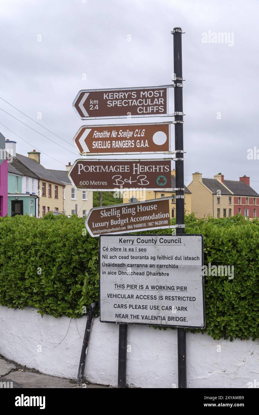 Vari cartelli forniscono indicazioni e indizi in una strada di case colorate e cieli grigi, West Coast, Portmagee, Iveragh Peninsula, County Kerry, Irelano Foto Stock