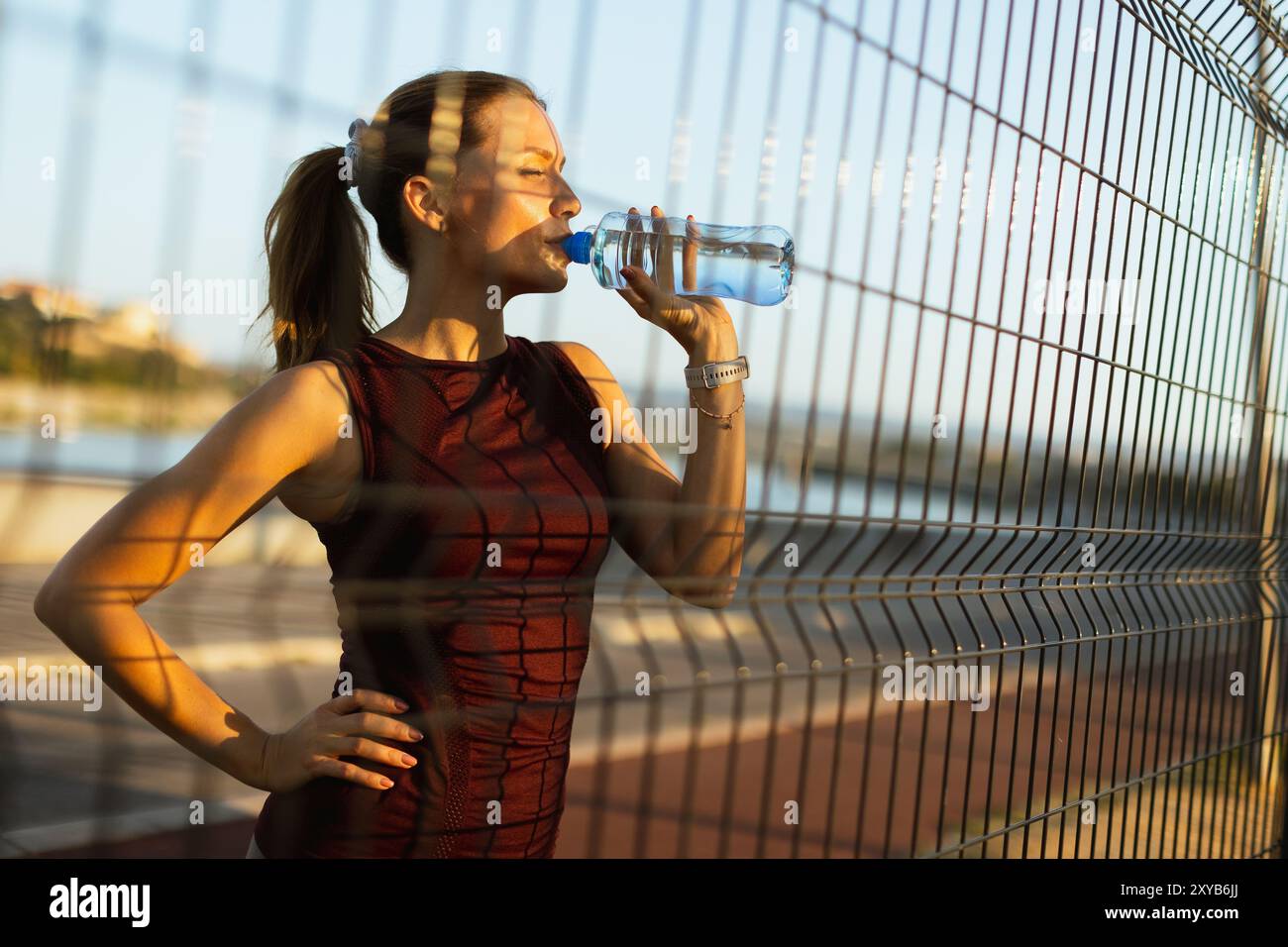 Con il sole che tramonta alle sue spalle, la donna si idrata dopo un allenamento all'aperto rinvigorente, prendendosi un momento per apprezzare la serena atmosfera lungo il fiume Foto Stock