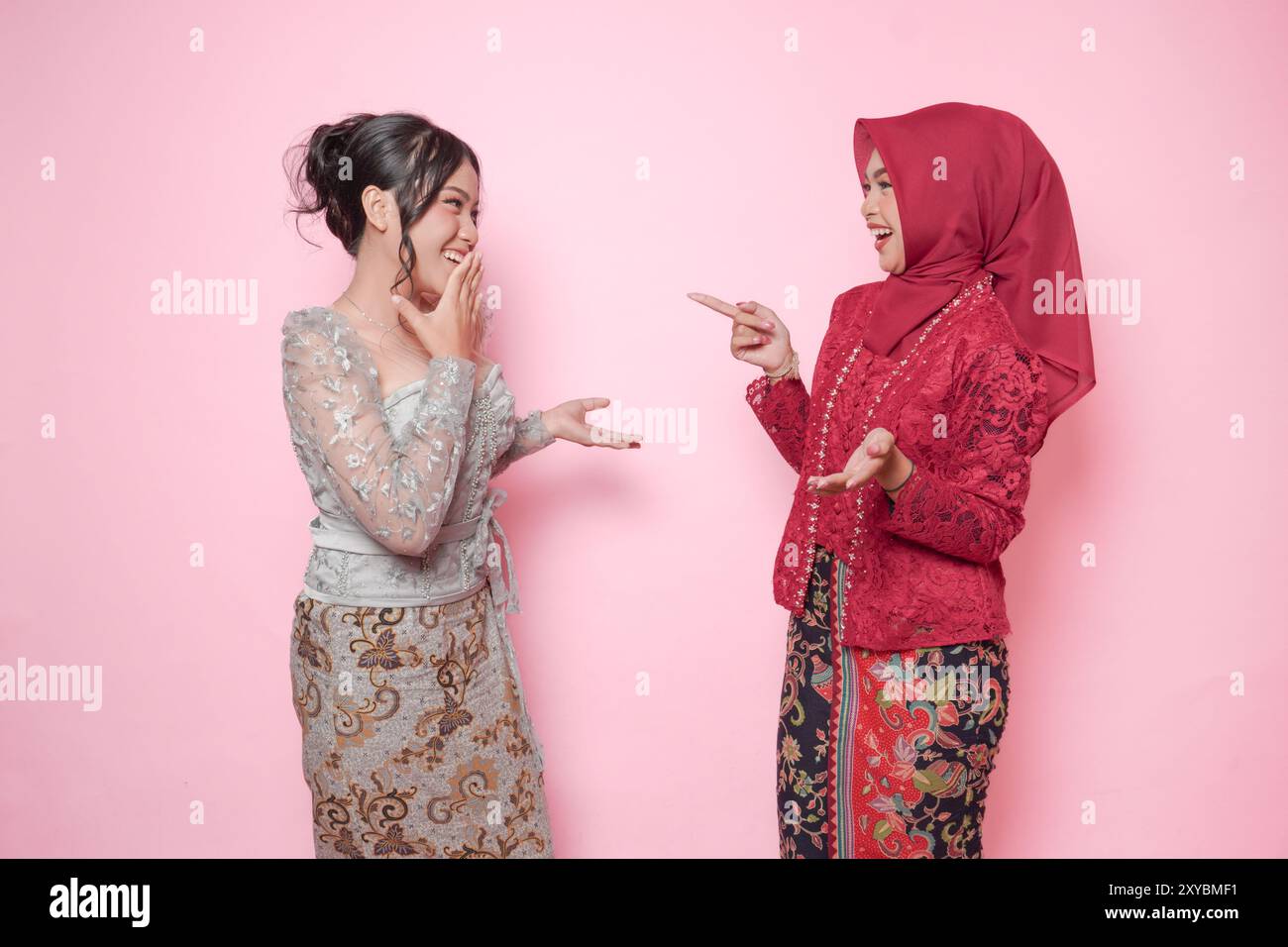 Due donne asiatiche come amici intimi che indossano kebaya moderna che fanno una divertente conversazione, isolata su sfondo rosa. Foto Stock