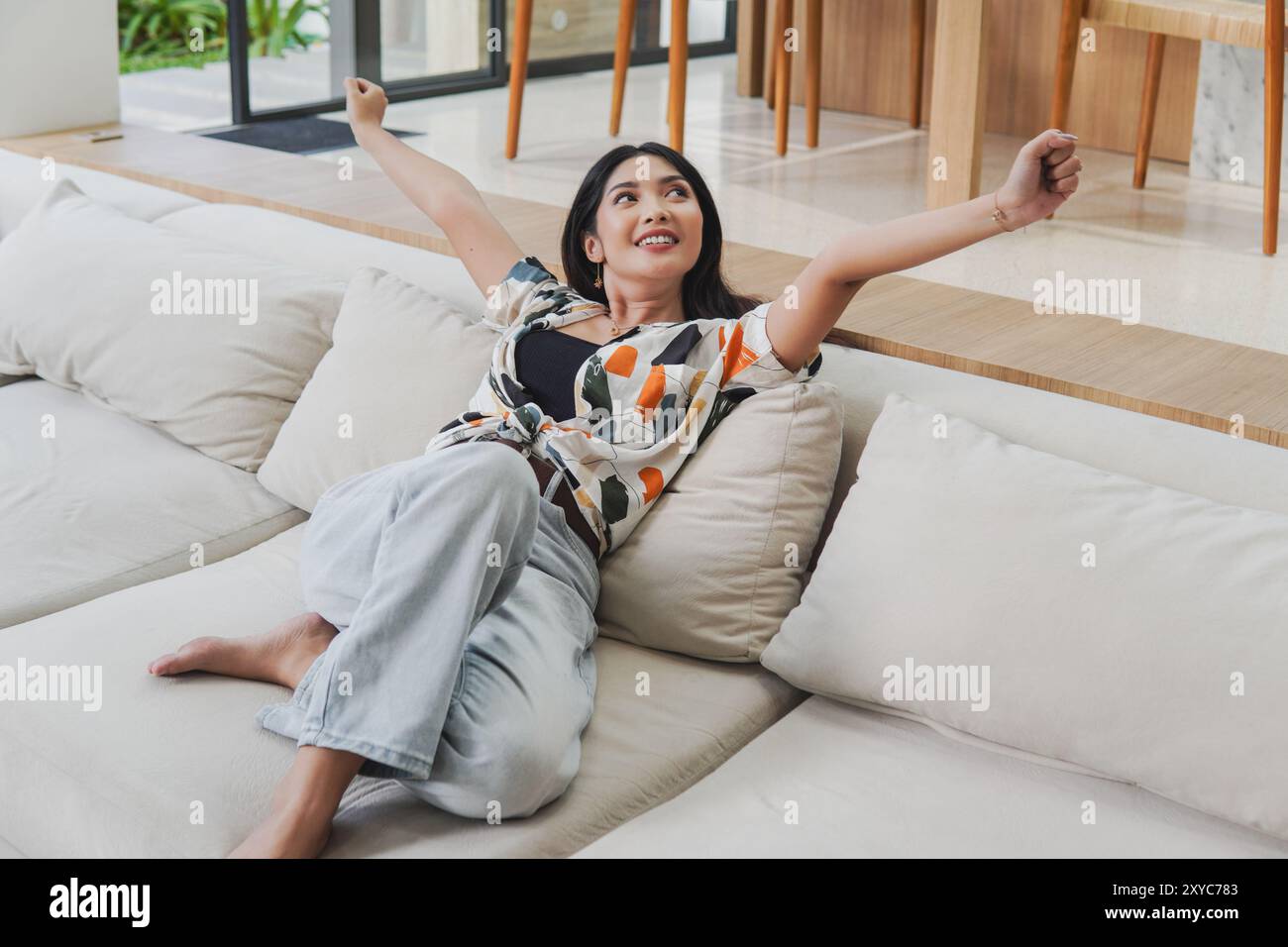 Bella giovane donna asiatica è seduta su un divano con le braccia alzate in aria, si sente felice e si diverte in vacanza. Foto Stock