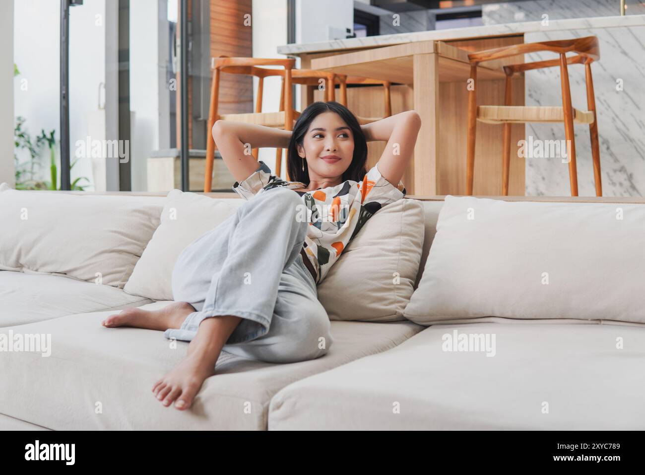 Bella giovane donna asiatica è seduta su un divano con le braccia alzate in aria, si sente felice e si diverte in vacanza. Foto Stock