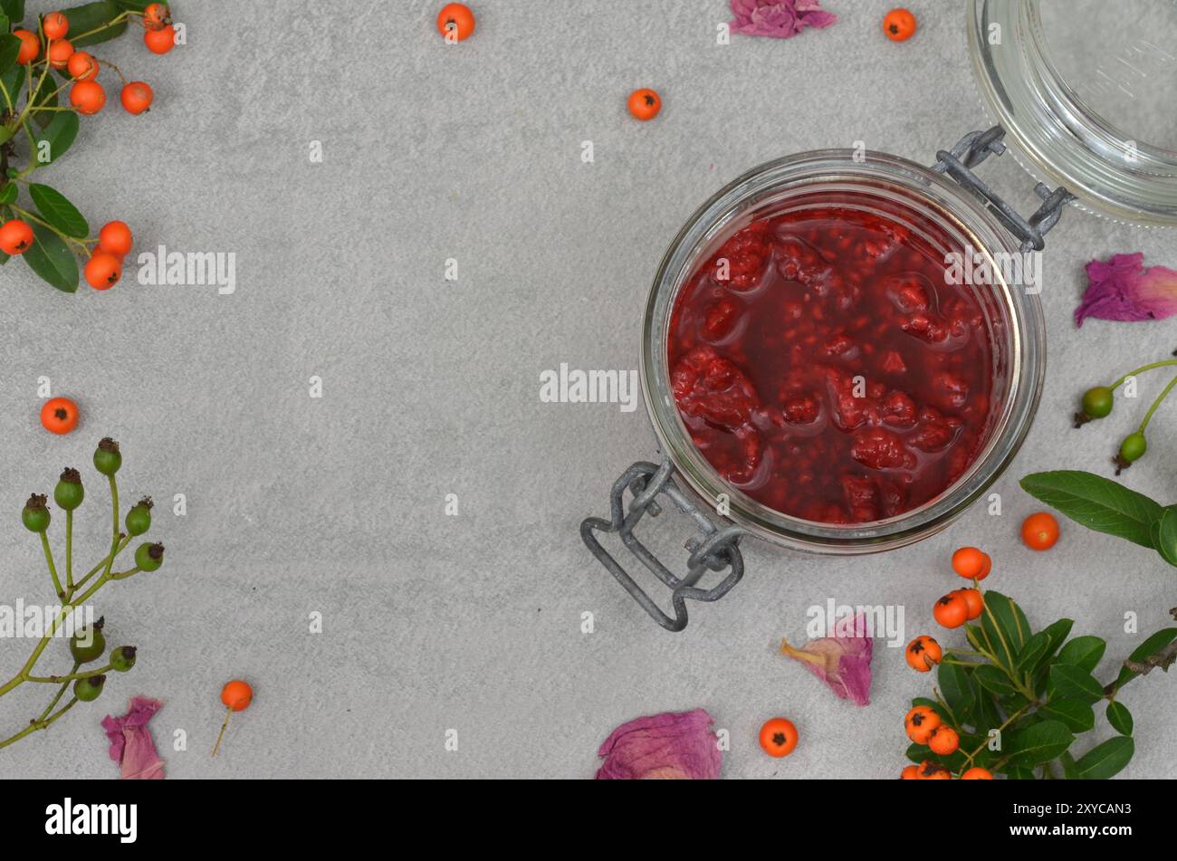 Marmellata di lamponi in vaso, decorata con foglie, ramoscelli, frutti di bosco, petali di fiori con spazio vuoto per riempire il contenuto, vista dall'alto. Sfondo grigio Foto Stock