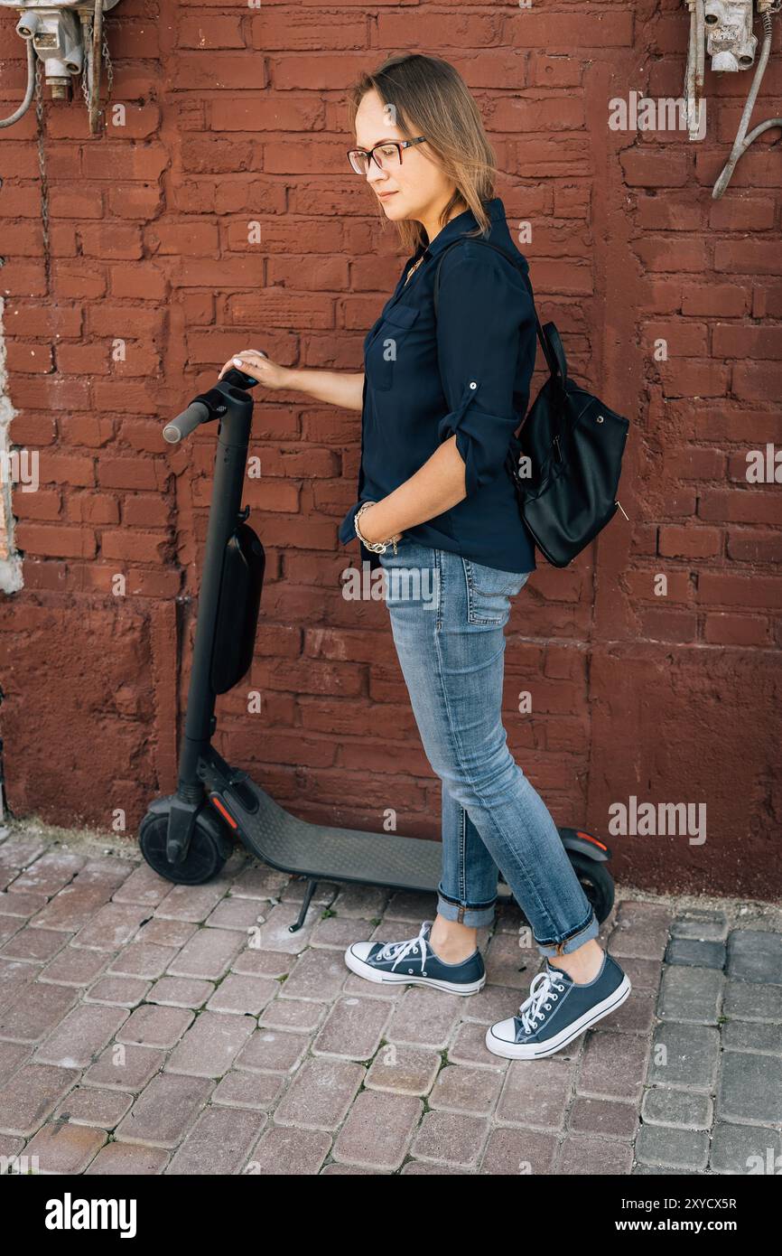 Una donna in sneakers alla moda cavalca uno scooter elettrico sulla parete di mattoni Foto Stock