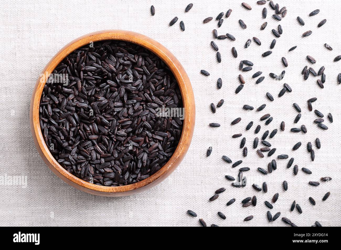 Riso nero, viola o riso proibito in una ciotola di legno su tessuto di lino. Oryza sativa, riso coltivato in Asia, cereali di colore nero intenso. Foto Stock