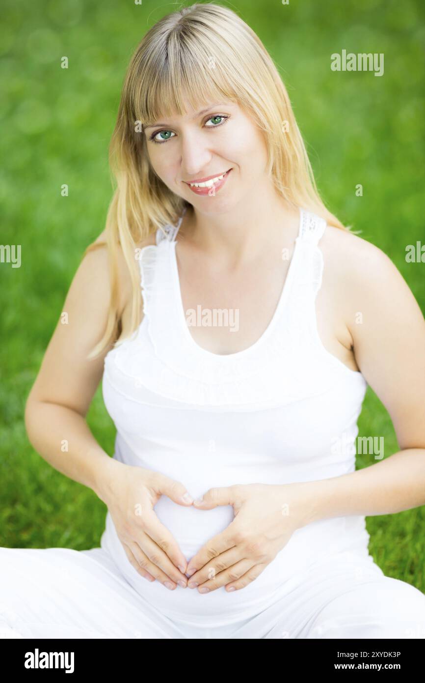 Felice sorridente donna incinta che tiene le mani sulla pancia a forma di cuore Foto Stock