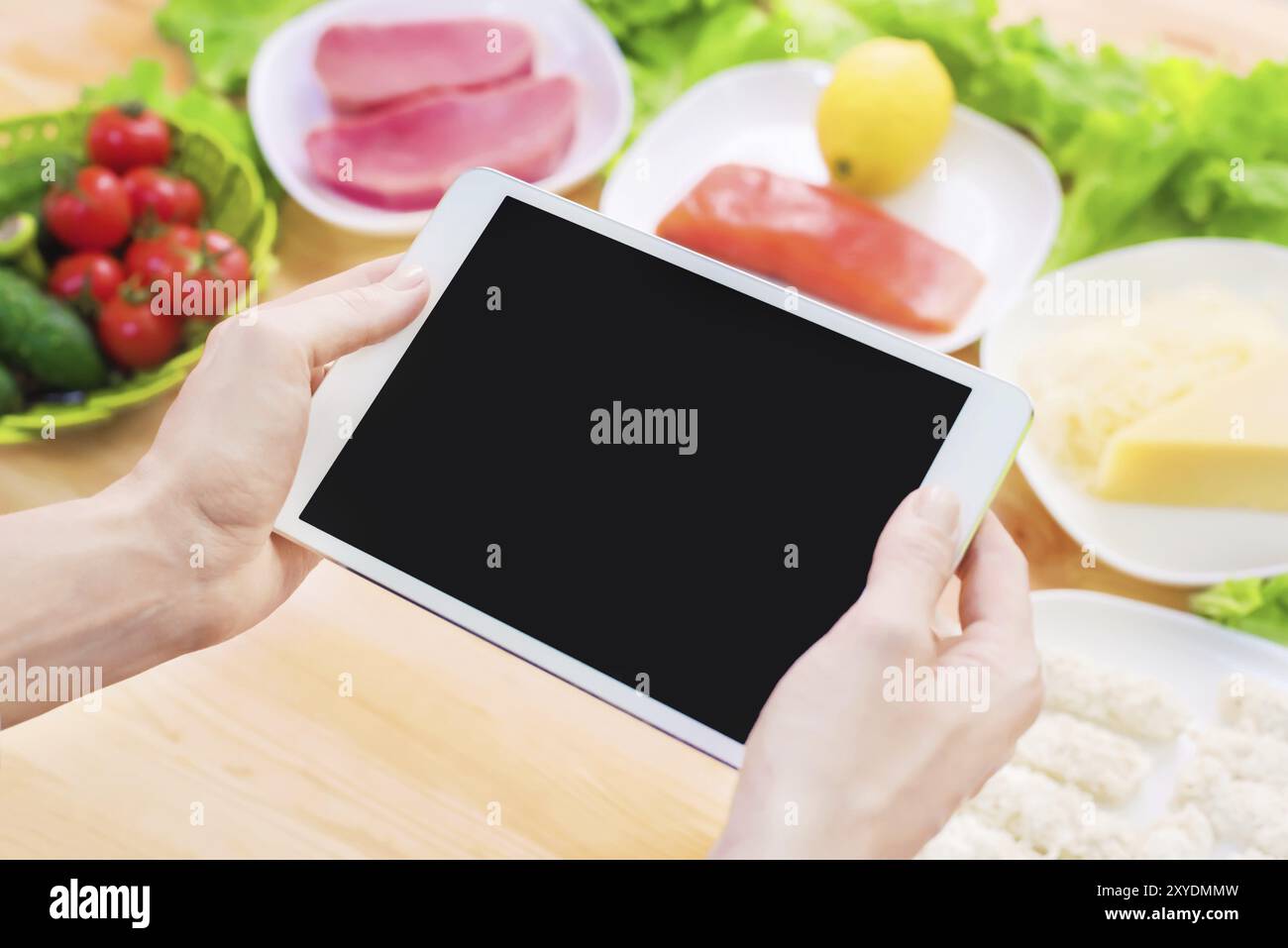 Vista dall'alto ravvicinata di mani femminili che tengono in mano un tablet computer con uno schermo vuoto circondato da cibo sano su un tavolo da cucina di casa. Pezzo da infilare Foto Stock