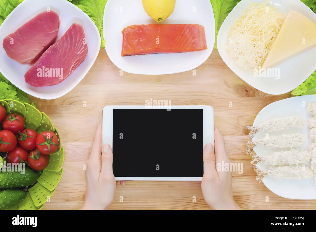 Vista dall'alto ravvicinata di mani femminili che tengono in mano un tablet computer con uno schermo vuoto circondato da cibo sano su un tavolo da cucina di casa. Pezzo da infilare Foto Stock