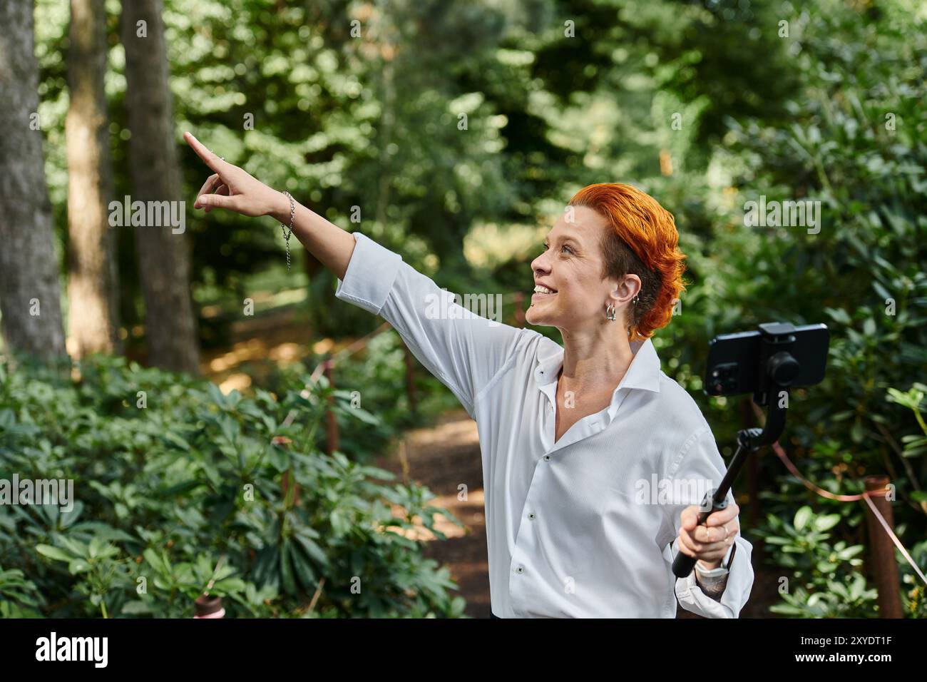 Una donna con i capelli rossi sorride e filma con un bastoncino per selfie in un'area boscosa del campus. Foto Stock