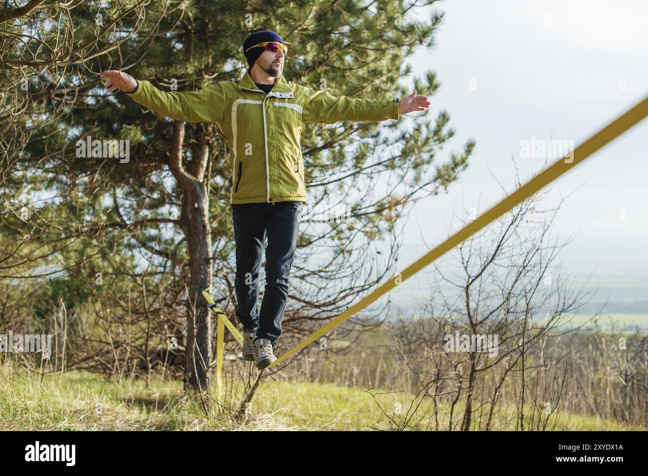 Un uomo, invecchiato con la barba e con gli occhiali da sole, si bilancia su una linea lenta all'aria aperta tra due alberi al tramonto sul cielo blu di sfondo Foto Stock