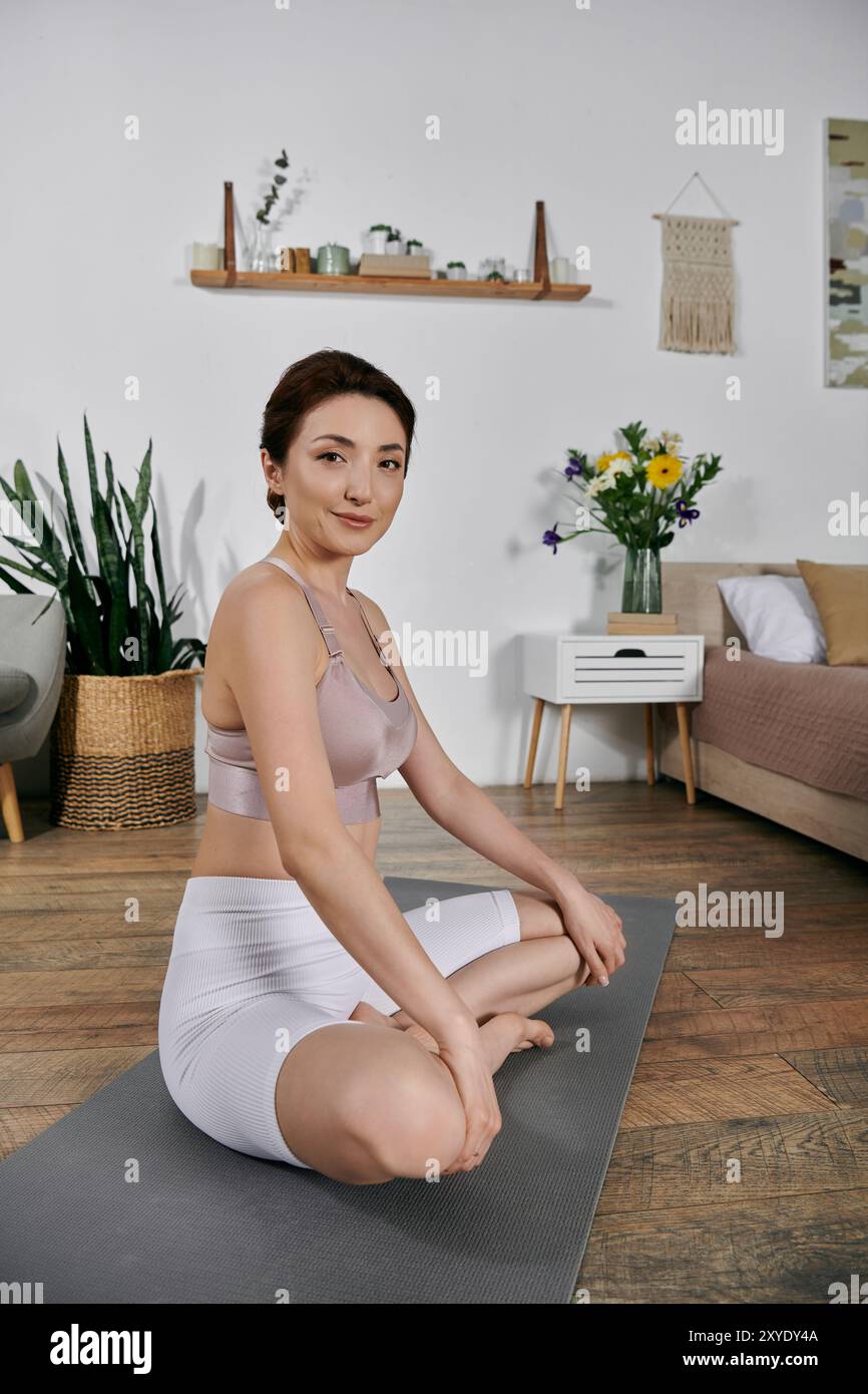 La donna asiatica in cima al raccolto e i pantaloncini meditano sul tappetino yoga in una moderna sala ben illuminata Foto Stock