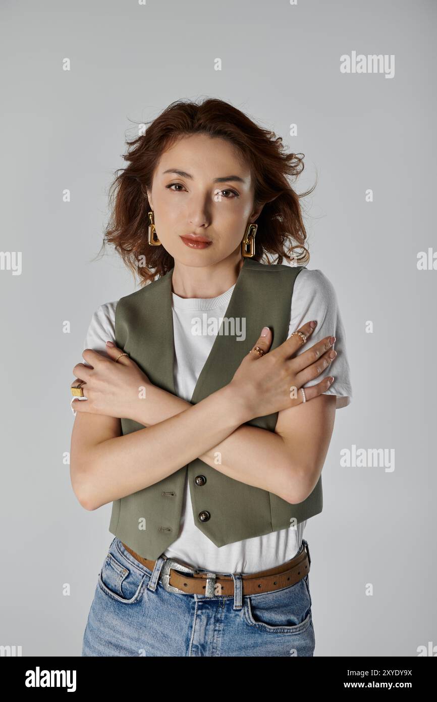 Una donna asiatica elegante e bella, che indossa un gilet e una sciarpa da collo, posa con le braccia incrociate su uno sfondo grigio. Foto Stock