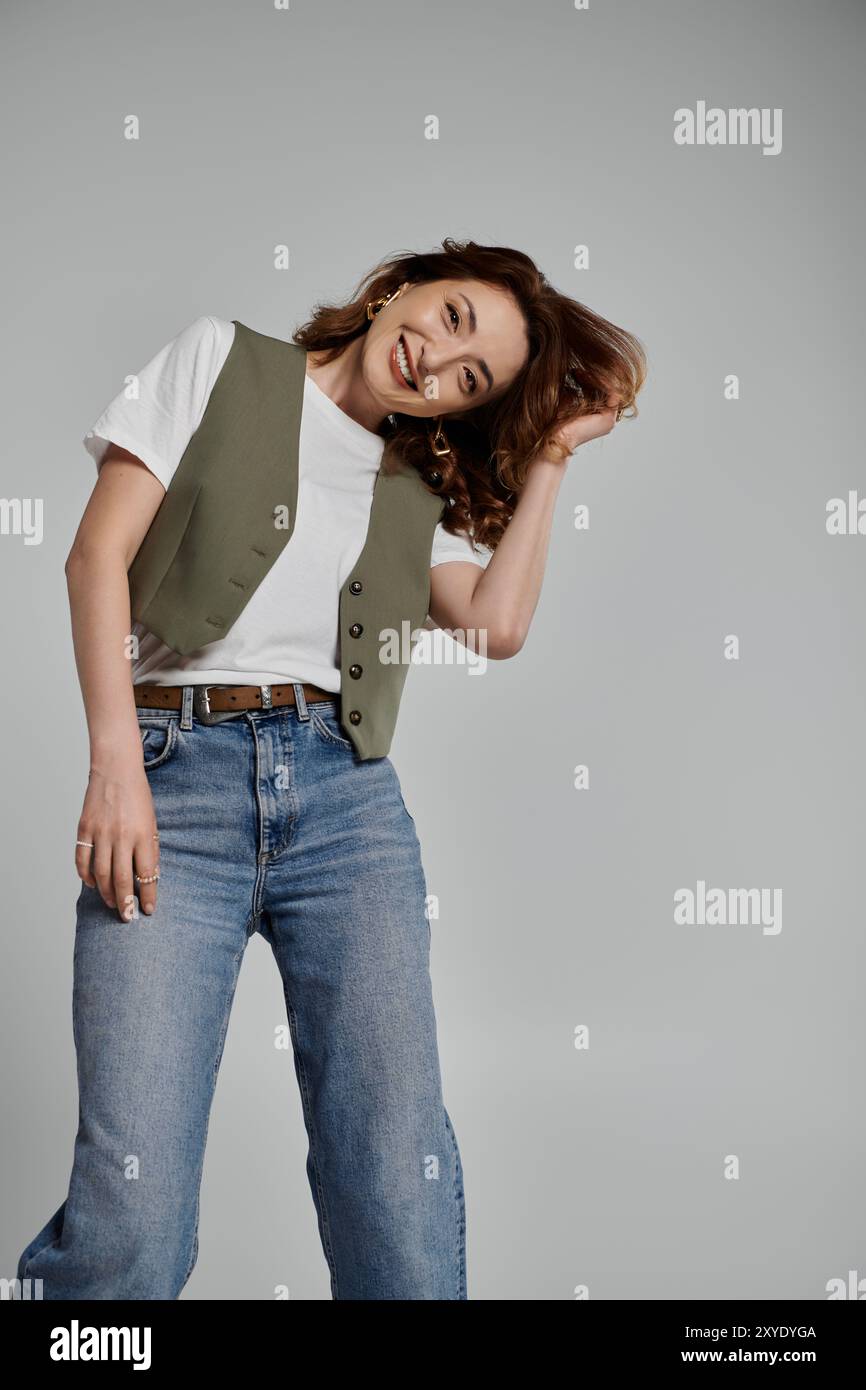Un'elegante donna asiatica che indossa un gilet verde, una t-shirt bianca e jeans si posa su uno sfondo grigio. Foto Stock