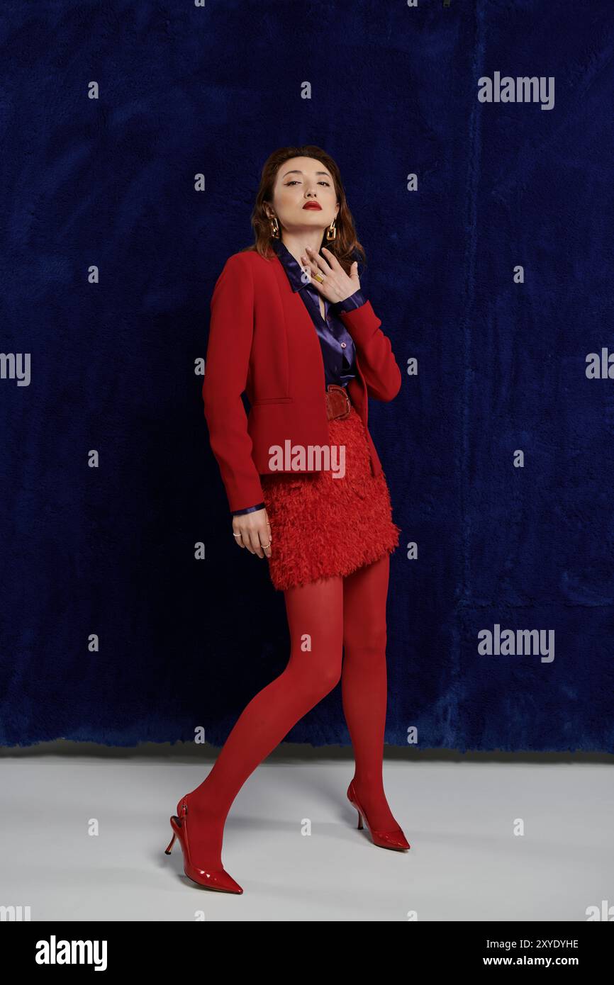 Una donna con vestito rosso, camicia di seta e calze poggia contro un muro blu. Foto Stock
