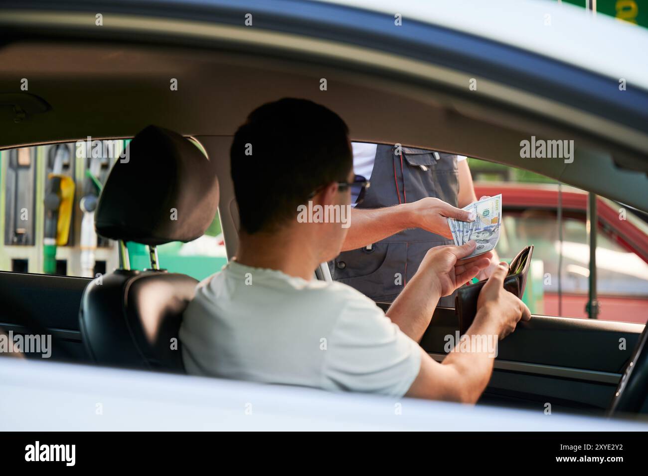 Giovane uomo che dà denaro all'operatore della stazione di servizio per il rifornimento dell'auto. Uomo che paga in contanti per il carburante. Concetto di spese di trasporto. Foto Stock