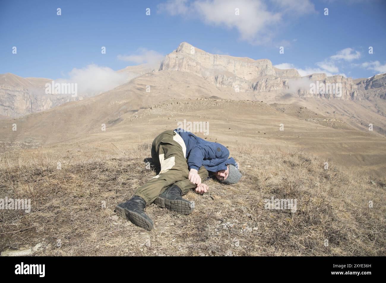 Un turista con un cappello e una barba giace per terra e si sente male in montagna. Una persona che ha perso conoscenza. Concetto di pericolo di incidente Foto Stock