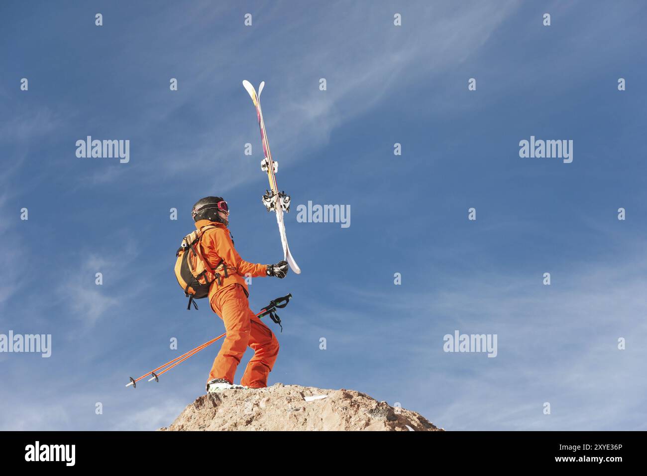 Uno sciatore-atleta in piedi sul bordo di un'alta roccia si bilancia con gli sci nelle mani contro il cielo blu e le nuvole di cirrus. Il concetto di sci in t Foto Stock