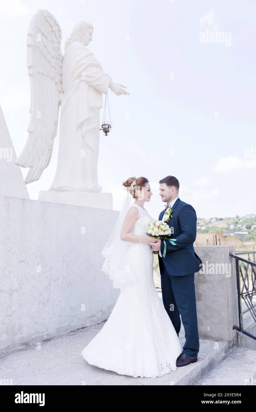 Ritratto di una bella coppia in luna di miele in un giorno di nozze con un bouquet in mano sullo sfondo di un monumento cristiano ortodosso con angeli. Foto Stock