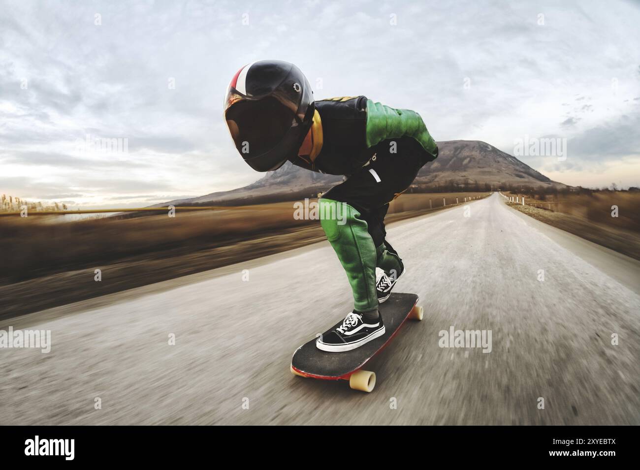 Un giovane con casco integrale e una tuta in pelle in uno speciale rack, cavalca un longboard sul tramonto afsaltu sulle montagne sullo sfondo e sul bellissimo sk Foto Stock Un giovane con casco integrale e una tuta in pelle in uno speciale rack, cavalca un longboard sul tramonto afsaltu sulle montagne sullo sfondo e sul bellissimo sk Foto Stock
