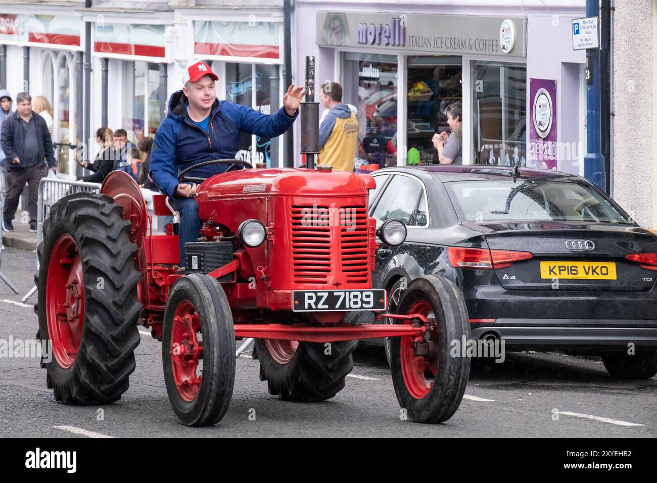 Uomo che indossa un cappello da baseball salutando gli spettatori, guidando un trattore rosso David Brown 25D d'epoca attraverso il centro città. Ballycastle, Regno Unito - 24 agosto 2024: Foto Stock