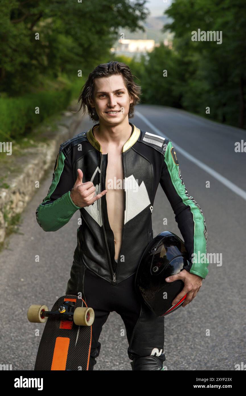 Un giovane uomo con un abito in pelle e capelli lunghi sta sull'asfalto con un casco tra le mani e il suo longboard sullo sfondo del verde e. Foto Stock Un giovane uomo con un abito in pelle e capelli lunghi sta sull'asfalto con un casco tra le mani e il suo longboard sullo sfondo del verde e. Foto Stock