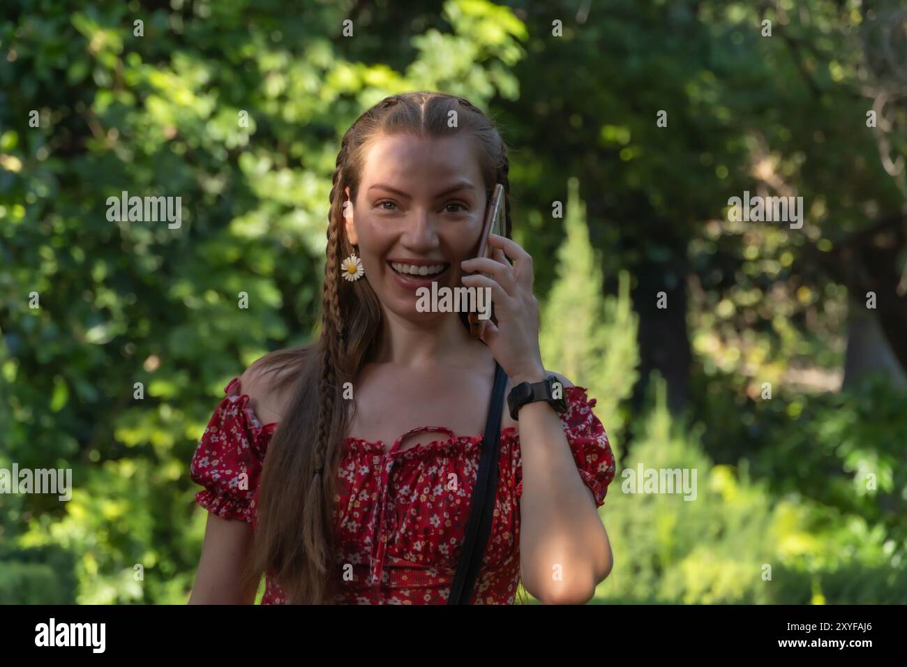 Una giovane rossa si gode una passeggiata spensierata in un giardino pieno di vita, chiacchierando al telefono. La luce del sole mette in risalto la sua serenità e gioia. Foto Stock