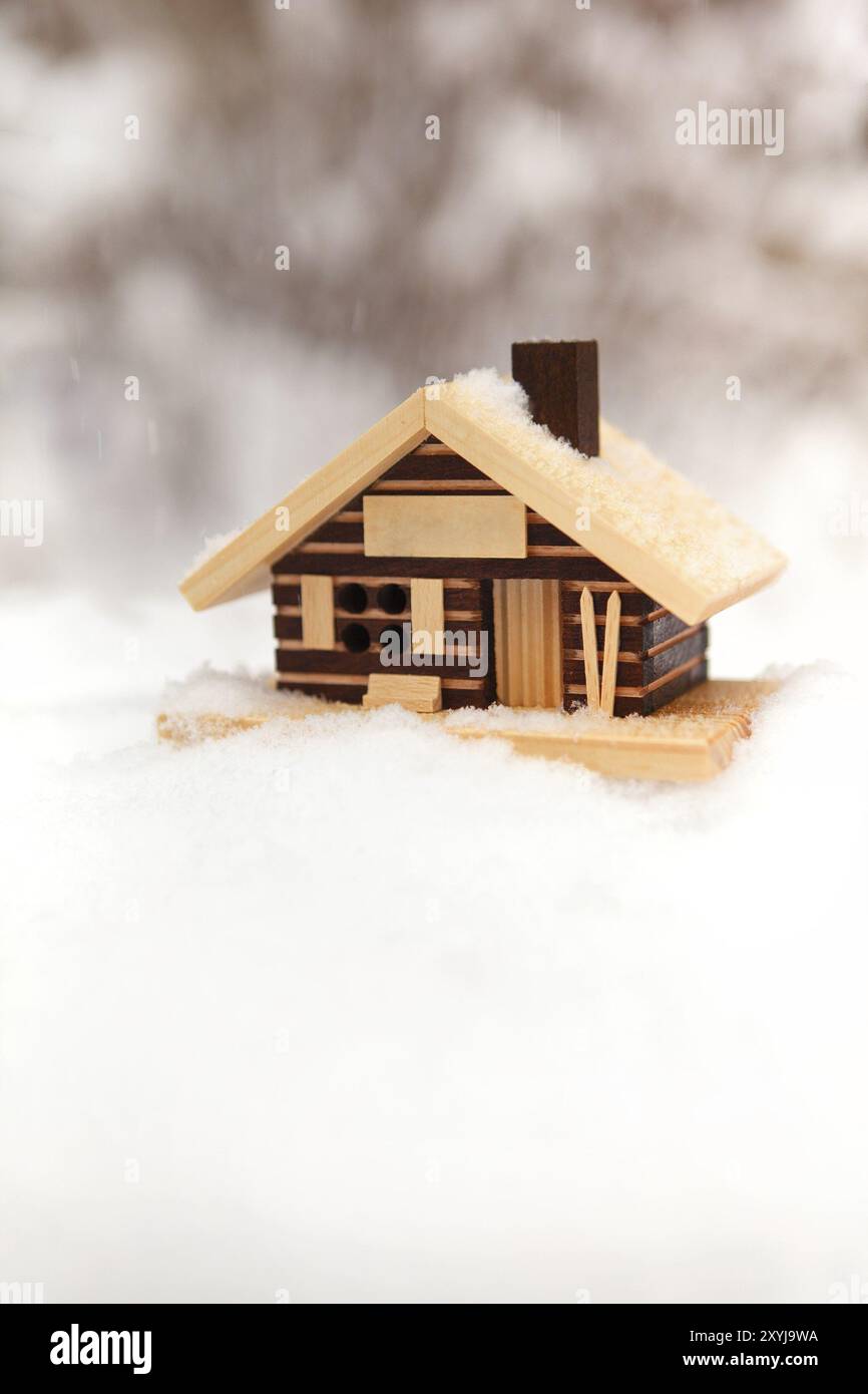 Piccolo modello di casa in legno tra neve con nevicata e sfondo offuscato foresta invernale Foto Stock