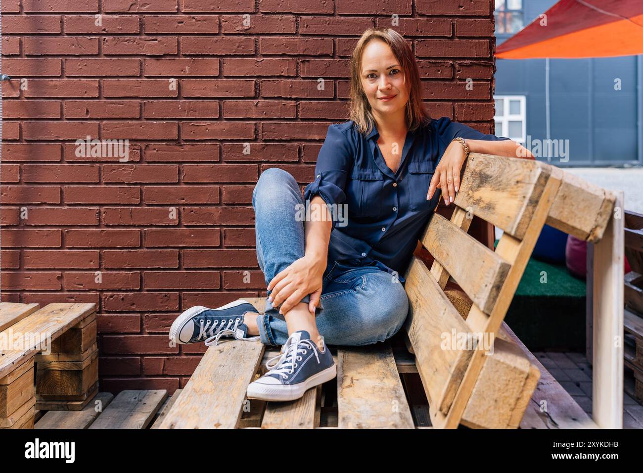 Donna seduta su pallet di legno contro una parete di mattoni all'aperto. Foto Stock