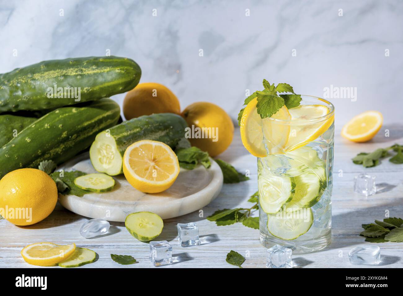 Un bicchiere di acqua cetriolo con fette di limone, foglie di menta e cubetti di ghiaccio, con cetrioli freschi e limoni su un tavolo accanto Foto Stock