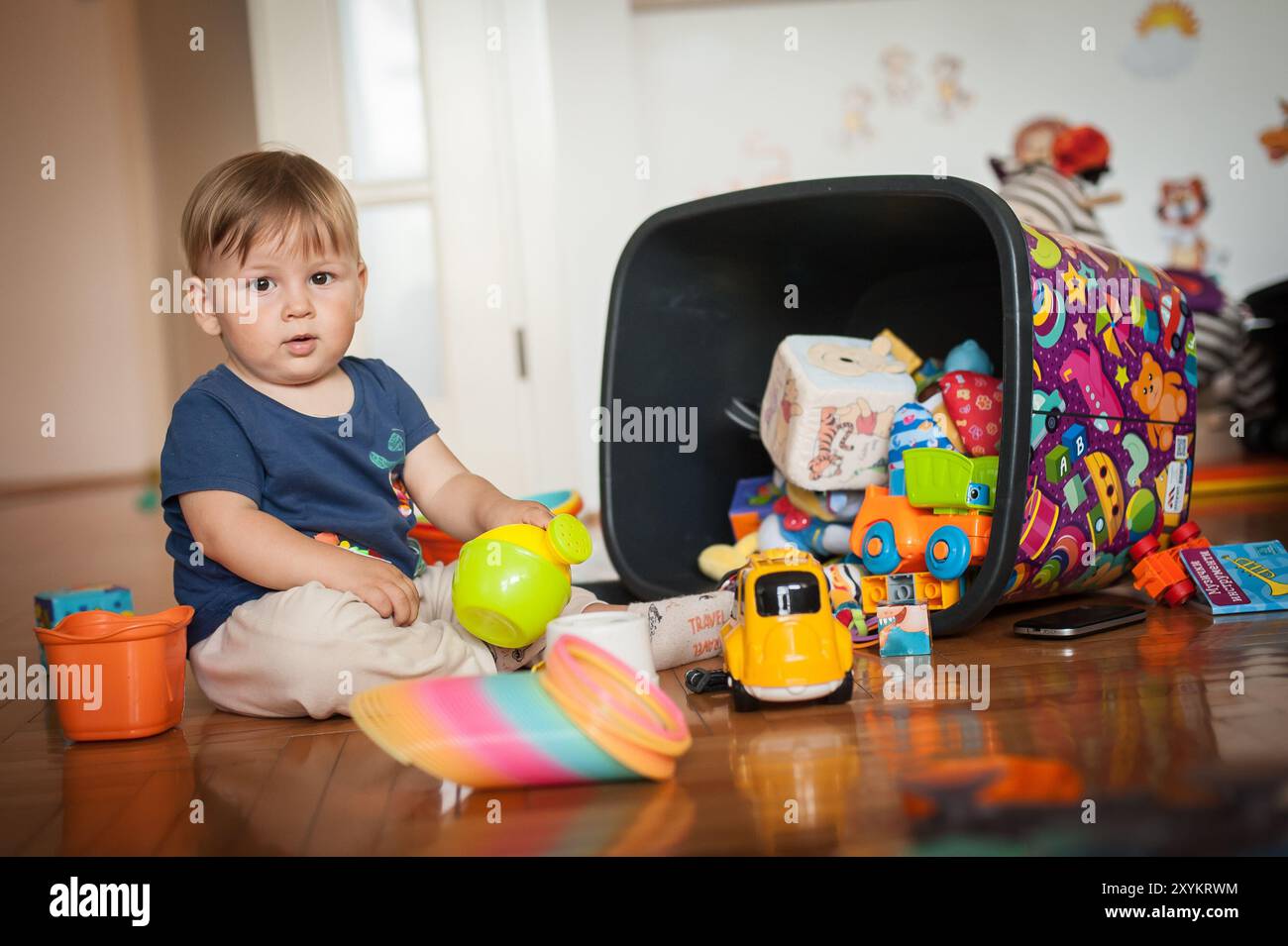 Un bambino si siede sul pavimento in una vivace sala giochi, scoprendo un tesoro di giocattoli fuoriusciti da un bidone rovesciato, creando un gioioso pasticcio Foto Stock