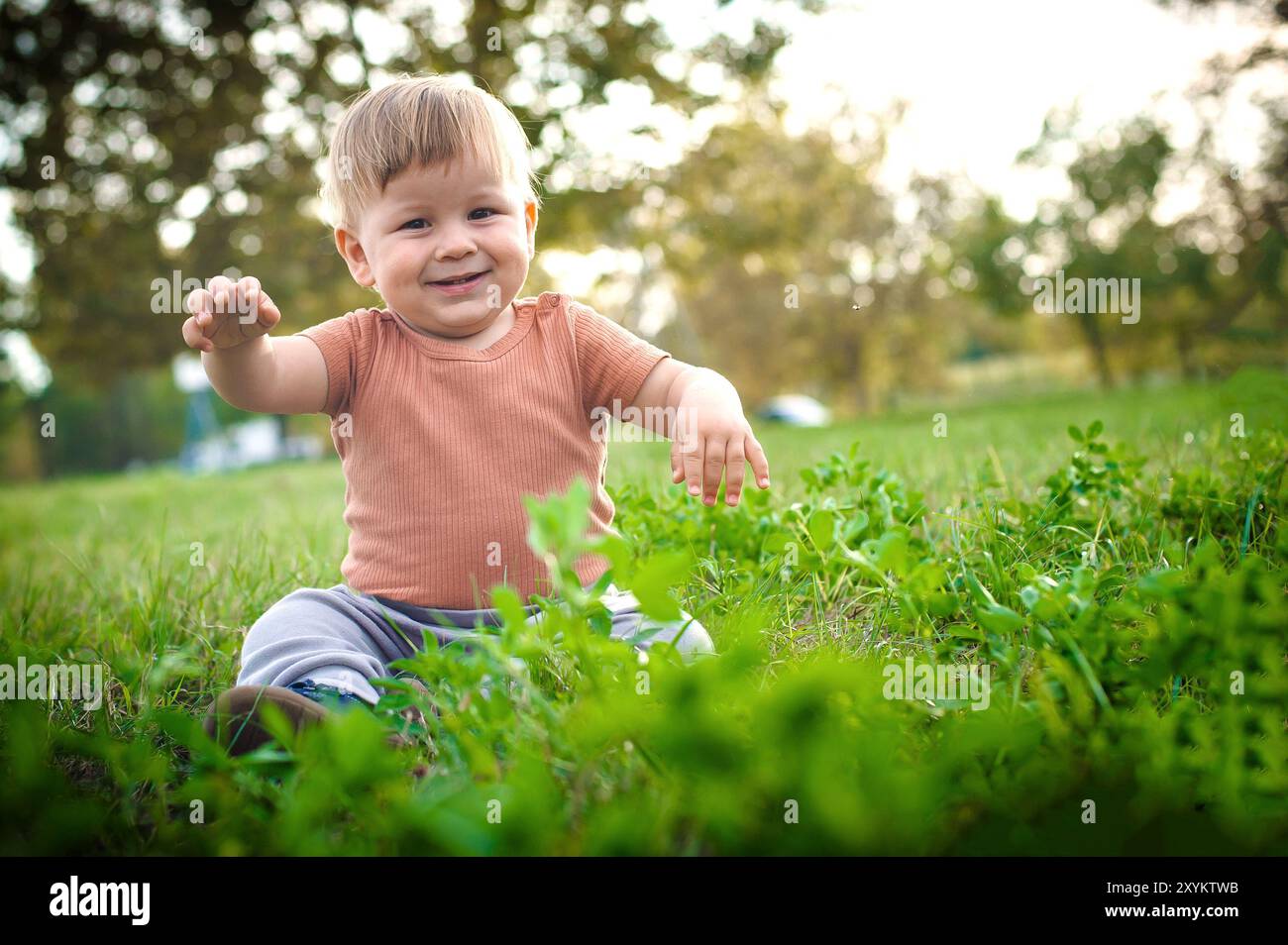 Un bambino allegro siede su una lussureggiante erba verde in un parco vivace, raggiungendo giocosamente la luce del sole bagna i dintorni, mostrando la sua meraviglia e j Foto Stock