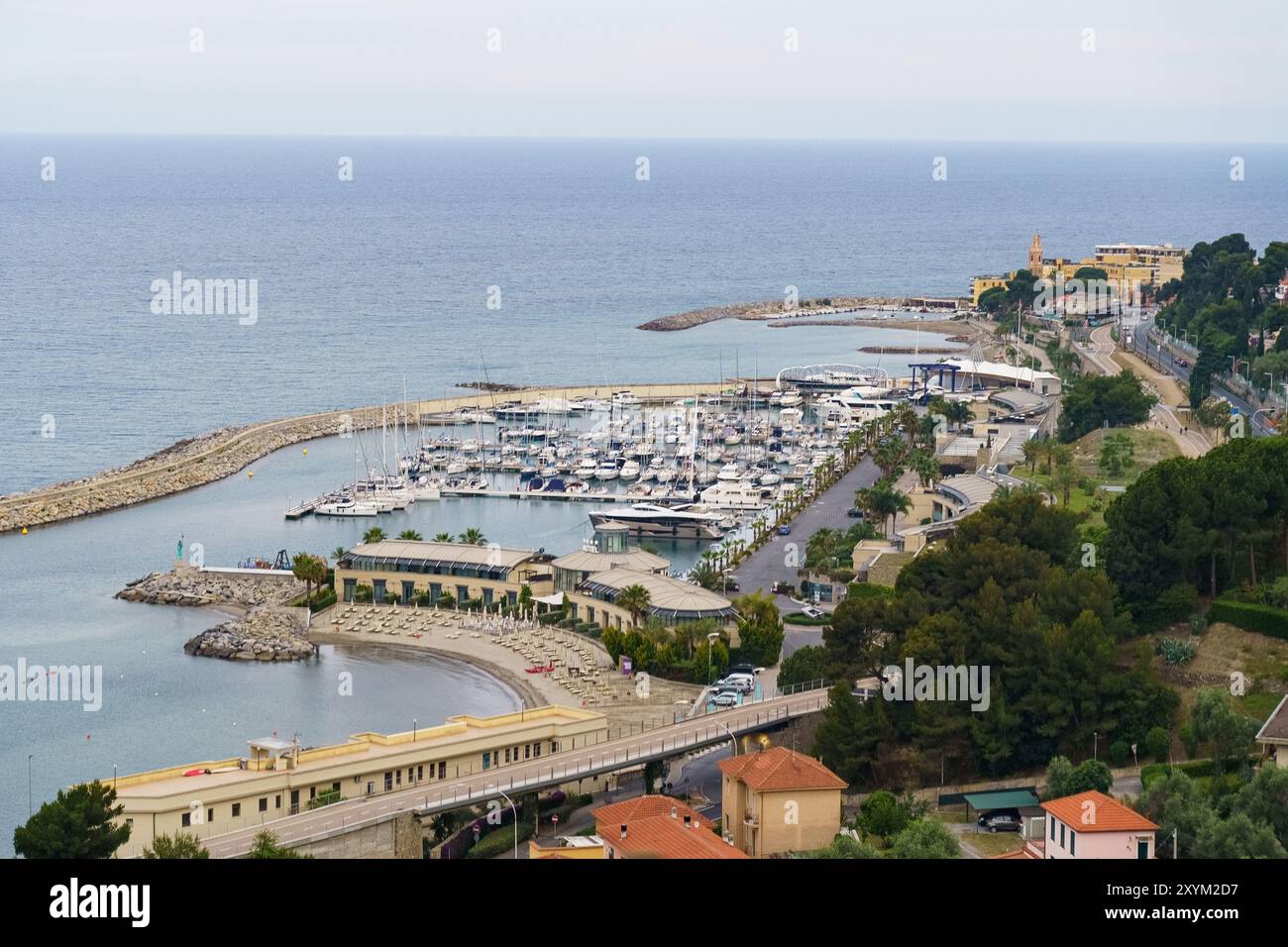 Un pittoresco porto nel Mar Mediterraneo con barche attraccate e una spiaggia vicina, circondato da vegetazione lussureggiante e affacciato su una città su una collina. Foto Stock