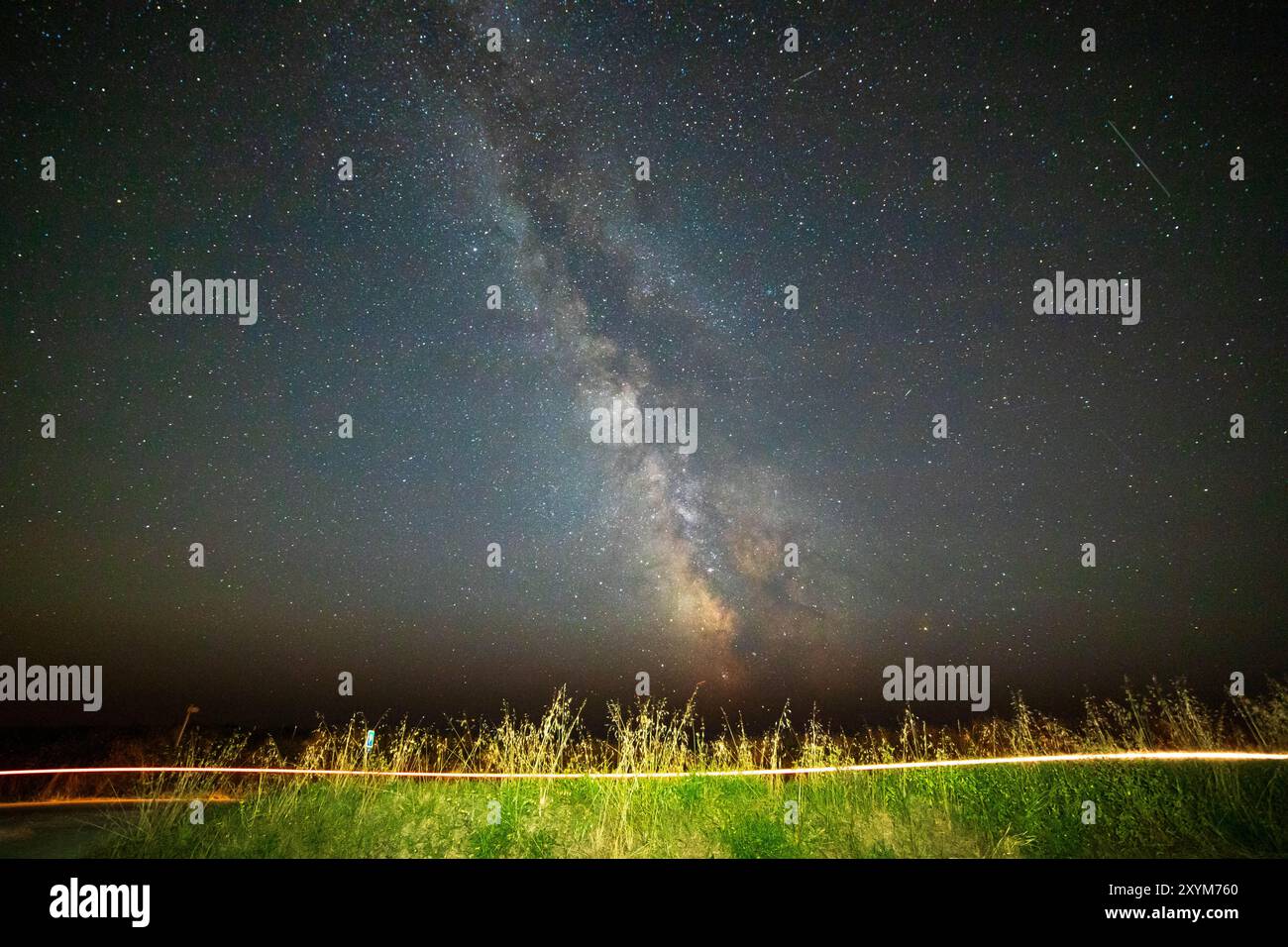 Cielo notturno con il nocciolo della via lattea sull'erba verde in un paesaggio di campagna, con una pista leggera lasciata da una moto. Brittany, Francia. Sfondo con poliziotto Foto Stock