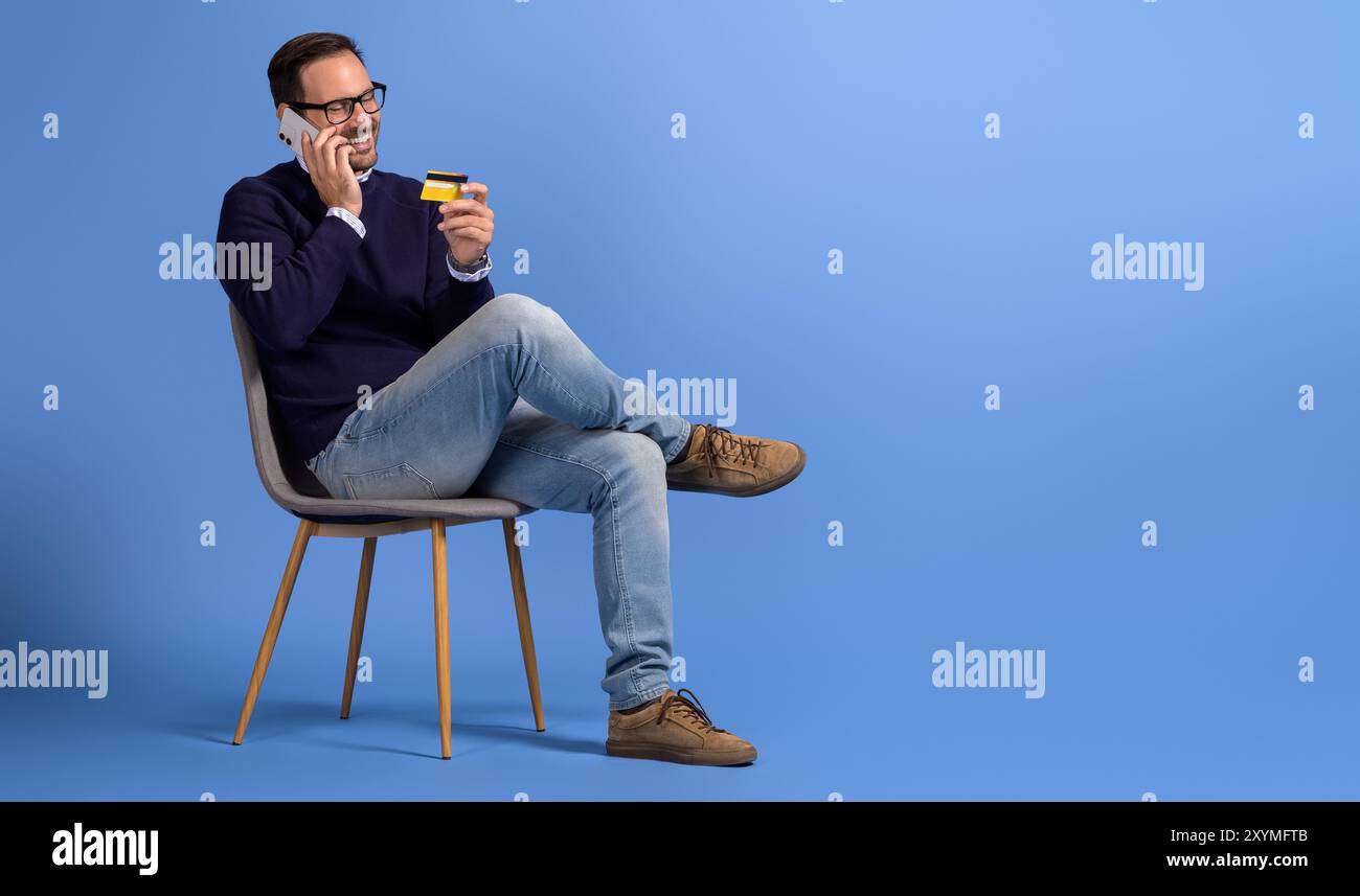 Giovane uomo d'affari felice che condivide i dettagli della carta di credito telefonando sulla sedia su sfondo blu Foto Stock