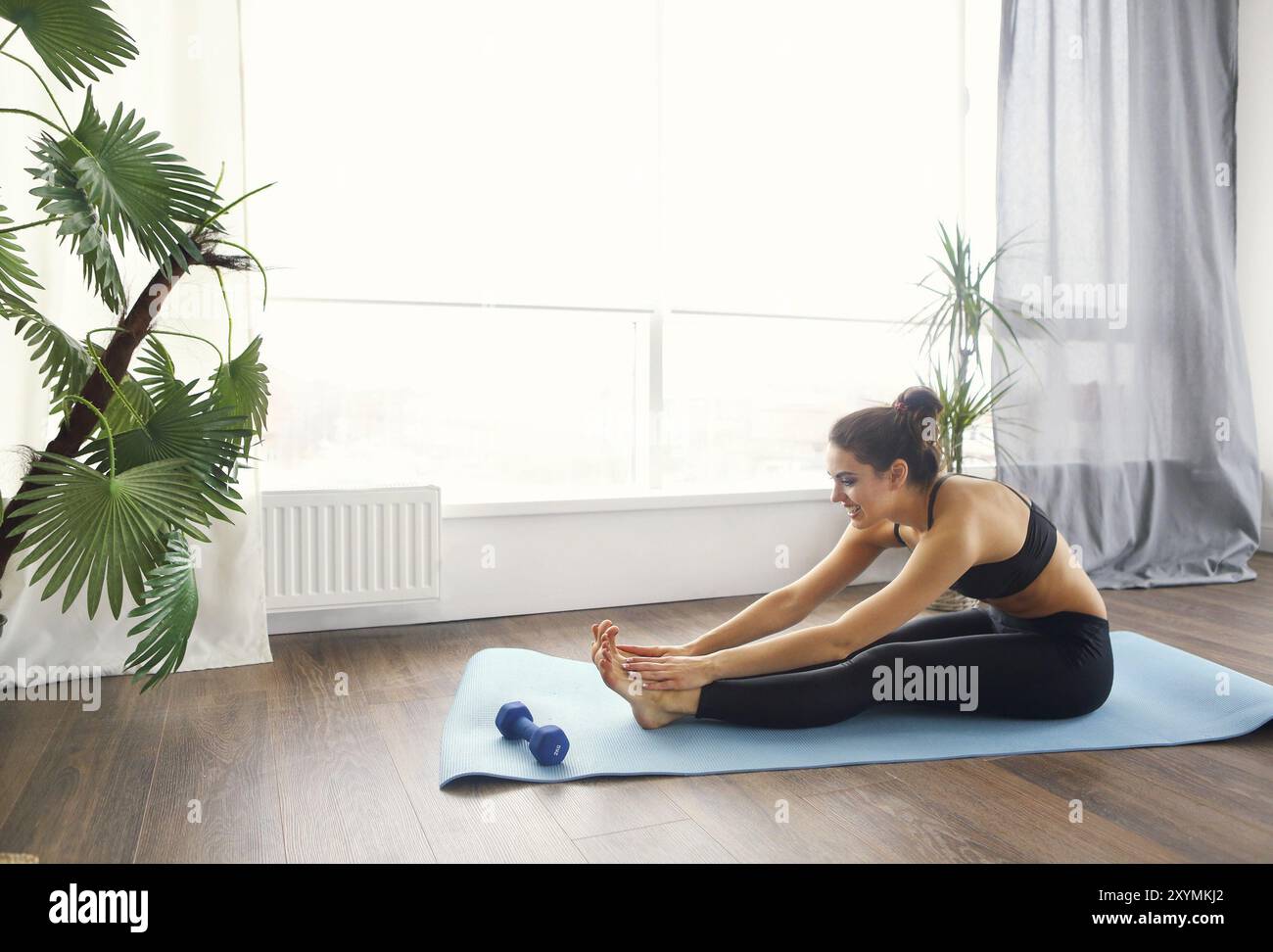 Donna sorridente in sportswear esercitando sul tappetino fitness e guardando la fotocamera in salotto Foto Stock