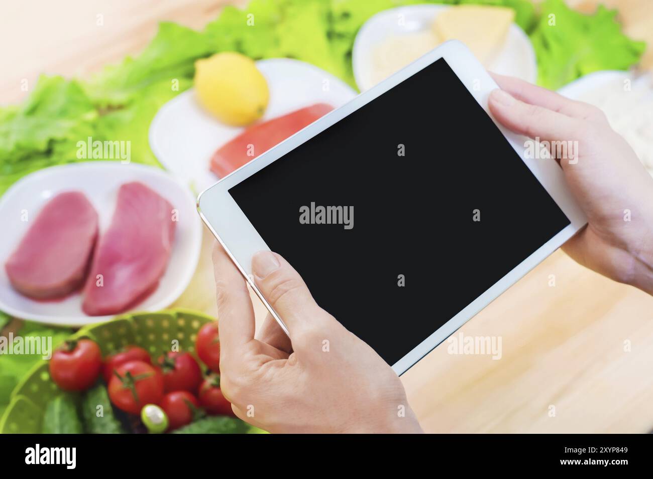 Vista dall'alto ravvicinata di mani femminili che tengono in mano un tablet computer con uno schermo vuoto circondato da cibo sano su un tavolo da cucina di casa. Pezzo da infilare Foto Stock