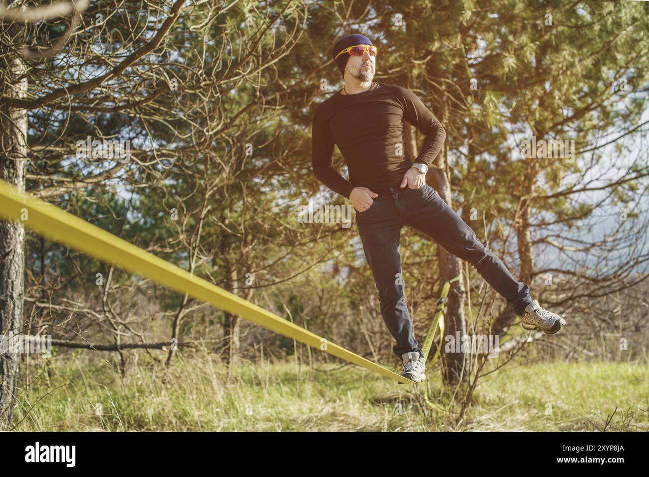 Un uomo, invecchiato con la barba e con gli occhiali da sole, si bilancia su una linea lenta all'aria aperta tra due alberi al tramonto sul cielo blu di sfondo Foto Stock