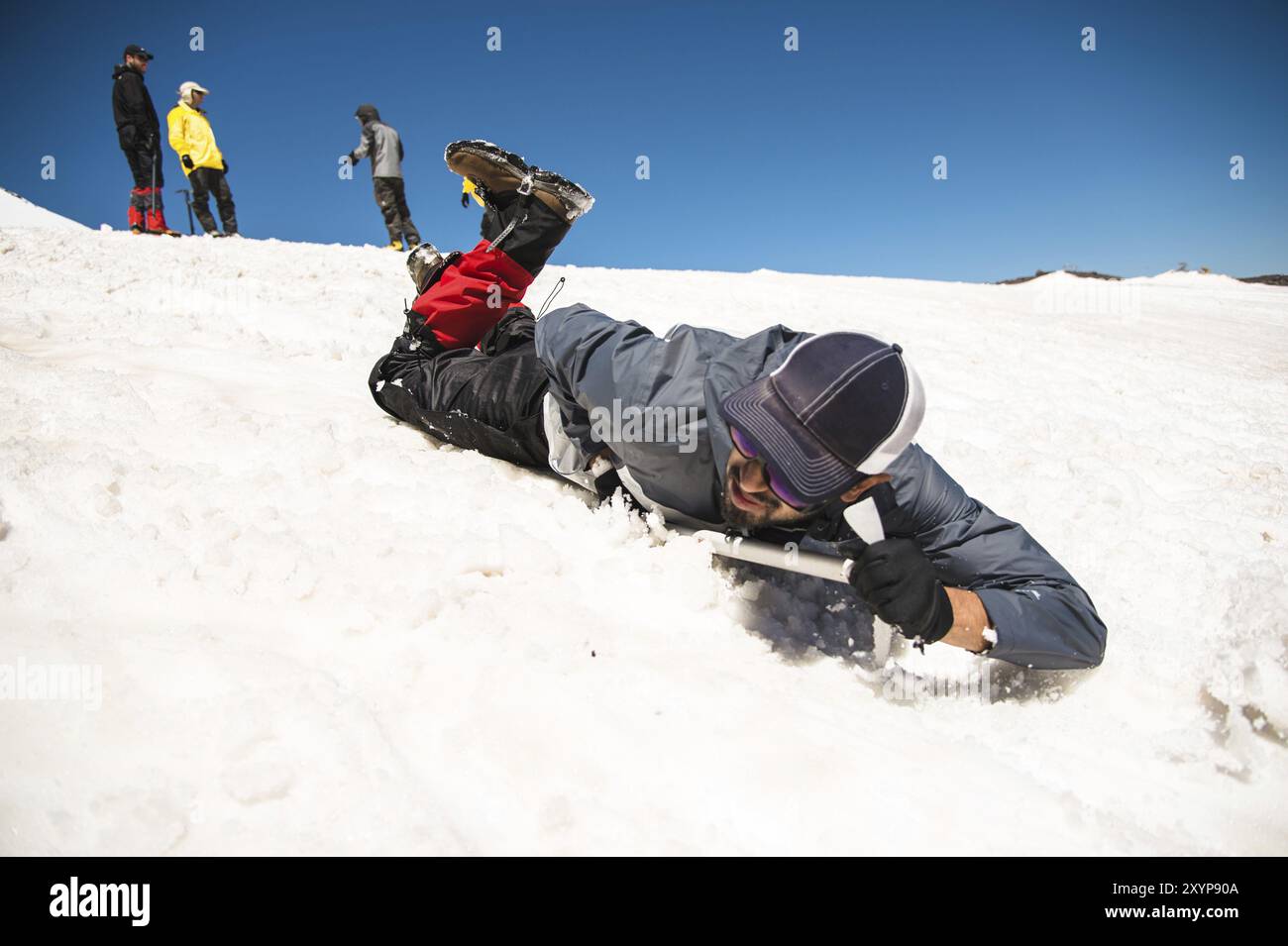 Allenamento per correggere lo scivolamento su un pendio o su un ghiacciaio con l'aiuto di un'ascia di ghiaccio. Un backpacker completamente attrezzato scivola sulla pancia lungo una pista innevata in th Foto Stock