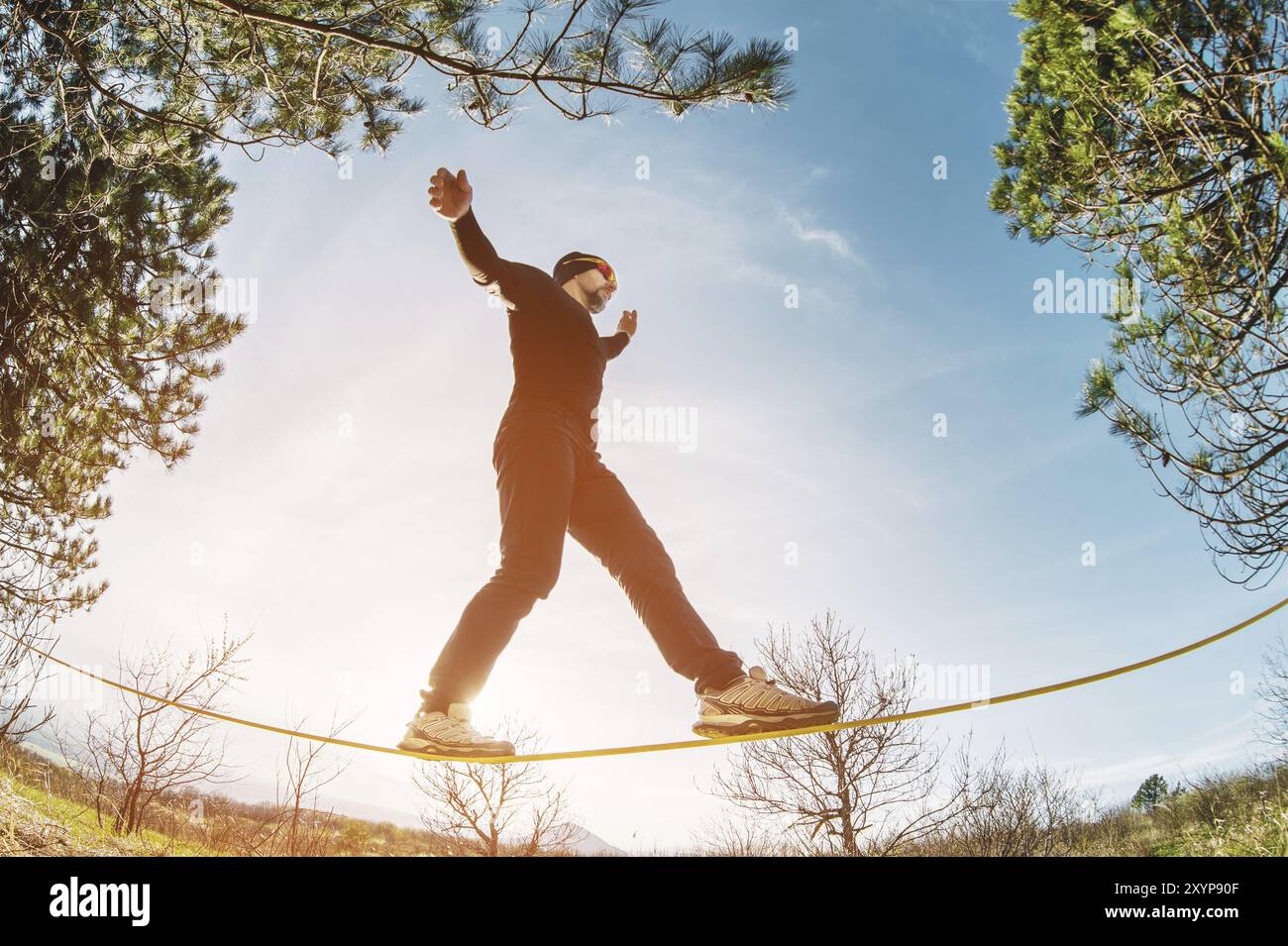 Un uomo, invecchiato con la barba e con gli occhiali da sole, si bilancia su una linea lenta all'aria aperta tra due alberi al tramonto sul cielo blu di sfondo Foto Stock