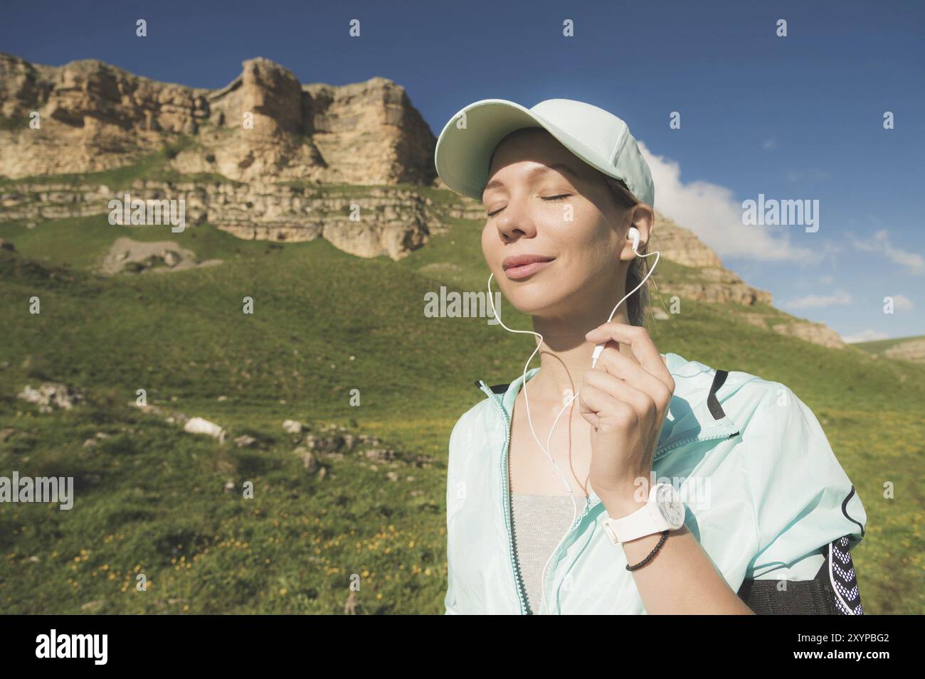 Donna atleta che chiude gli occhi ascoltando musica sulla natura. Ritratto di una bella ragazza che indossa auricolari e berretto da corsa. contro Foto Stock