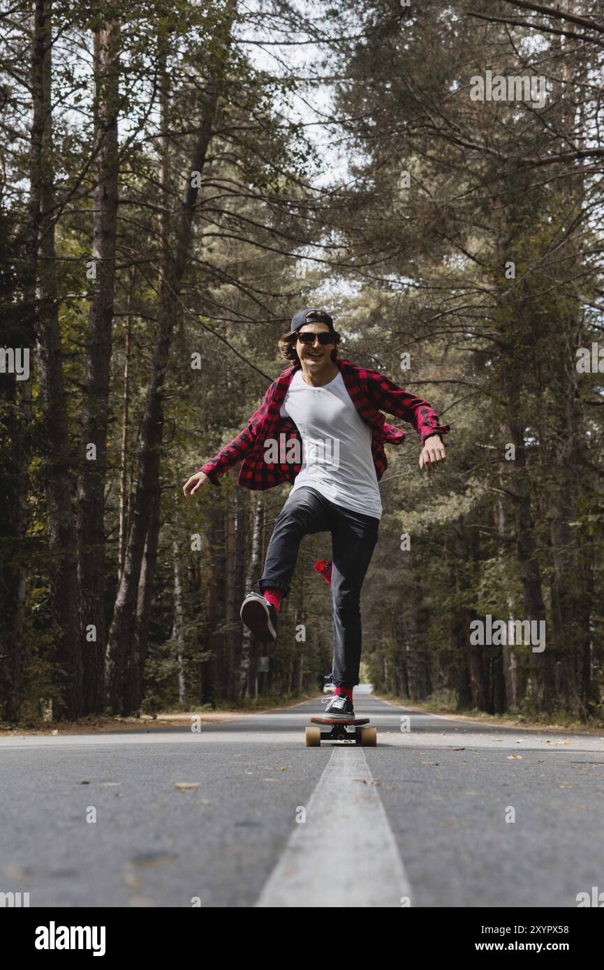 Un giovane hipster con cappello e camicia a quadri e occhiali da sole sta cavalcando il suo skateboard su una strada di campagna in una foresta autunnale. La fase del movimento di d Foto Stock
