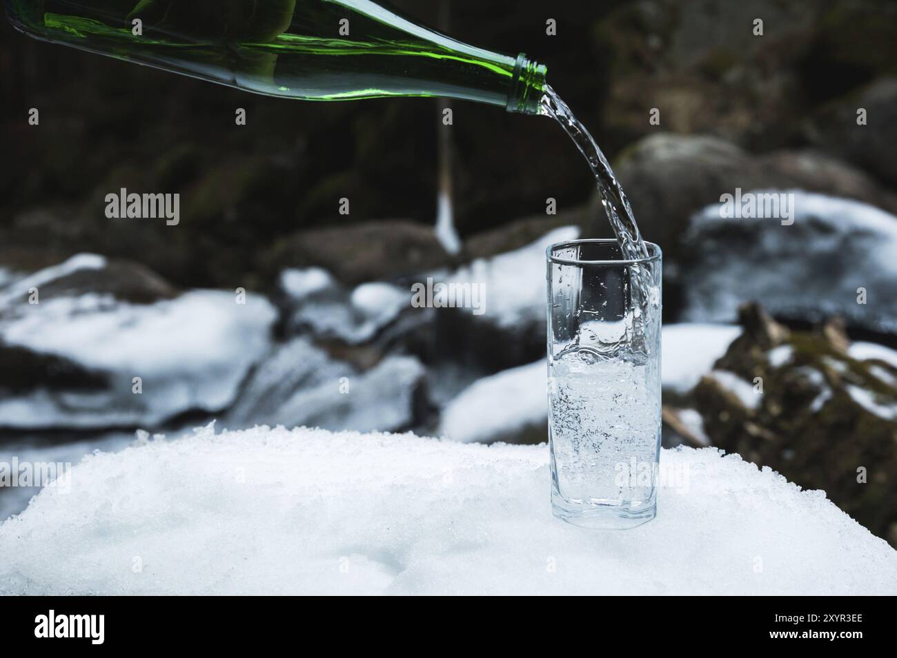 L'acqua minerale viene versata da una bottiglia verde in vetro in un bicchiere in vetro trasparente. Un bicchiere sta nella neve. Sullo sfondo di un inverno Foto Stock