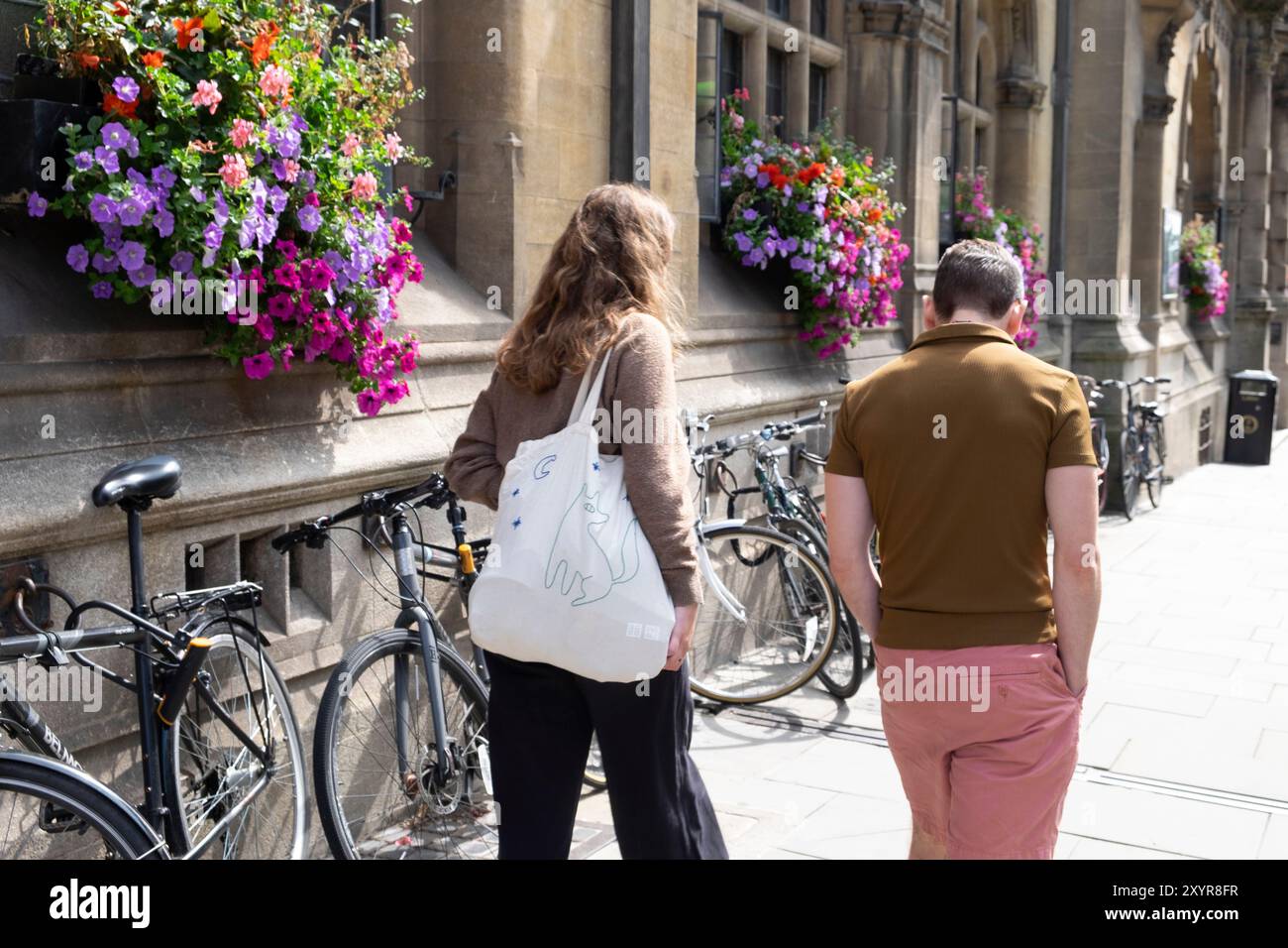 Vista posteriore di due persone uomo donna che cammina lungo la strada con bellissimi cesti appesi e biciclette appoggiate contro il muro dell'edificio nel centro di Oxford, Regno Unito Foto Stock