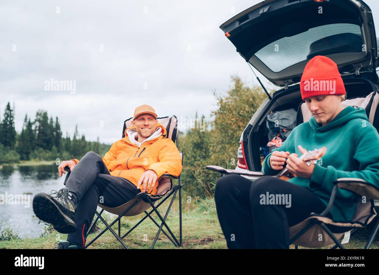 Tranquillo campeggio norvegese con padre e figlio sul lago. Si rilassano sulle sedie mentre il figlio gioca a ukulele circondati dalla natura, ridendo divertendosi Foto Stock