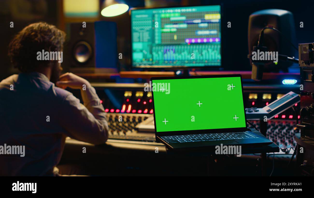 Il tecnico dell'audio regola il suono sulla console di mixaggio del registratore, con un computer portatile che mostra un display verde. Il produttore alza il volume dei brani in studio professionale, configurazione della sala di controllo. Telecamera A.. Foto Stock