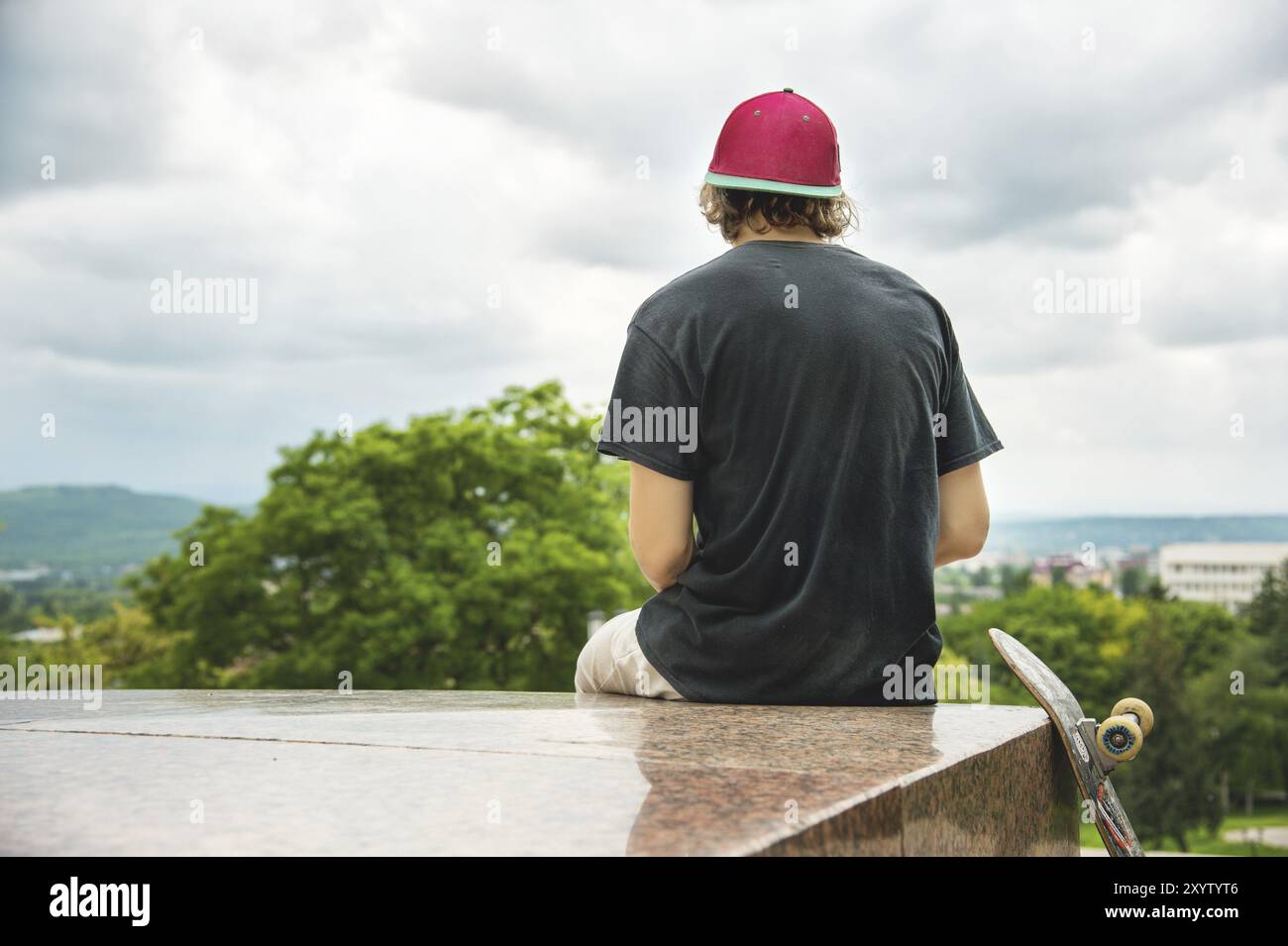 Lo skater dai capelli lunghi con un berretto e una maglietta si siede con la schiena alla telecamera e pensa accanto allo skateboard sullo sfondo del paesaggio Foto Stock