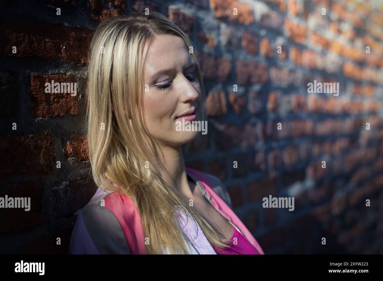 Giovane donna sognante appoggiata contro un muro di mattoni alla luce del sole basso Foto Stock