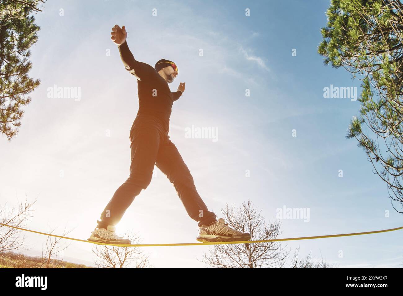 Un uomo, invecchiato con la barba e con gli occhiali da sole, si bilancia su una linea lenta all'aria aperta tra due alberi al tramonto sul cielo blu di sfondo Foto Stock