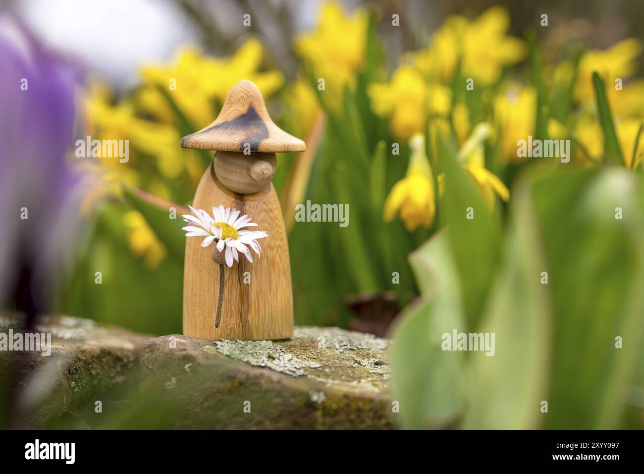 La figura di gnomi in legno con una margherita in mano si erge in un letto di fiori davanti a fiori gialli sfocati Foto Stock