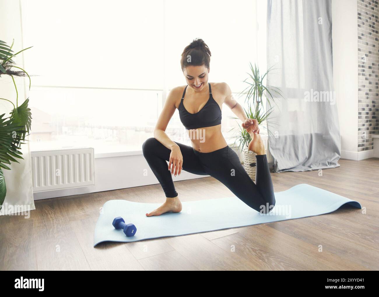 Donna sorridente in sportswear esercitando sul tappetino fitness e guardando la fotocamera in salotto Foto Stock